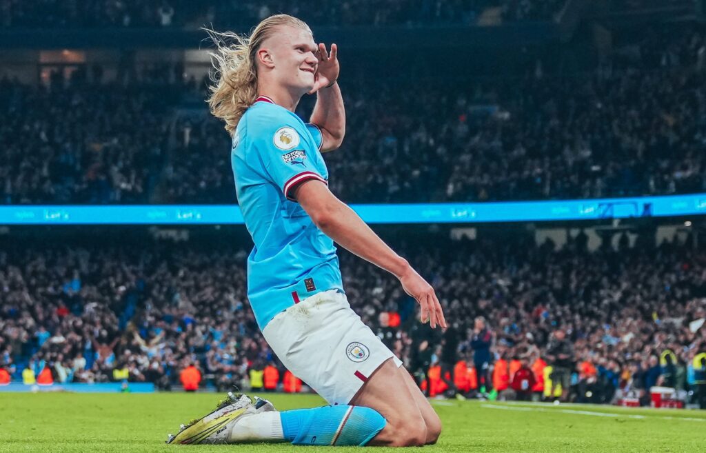 Haaland se torna o maior artilheiro da Premier League em uma edição - Jogador aparece ajoelhado no gramado. Ele veste uniforme azul com calção branco e está com cabelos loiros e soltos.