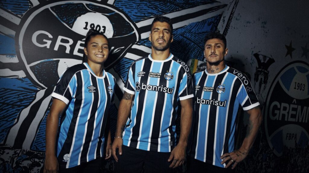 Nova camisa do Grêmio para 2023 é lançada; confira as fotos - jogadores do Grêmio posam para foto de divulgação da nova camisa do Grêmio, ao fundo uma parede com o escudo do Grêmio pintado.