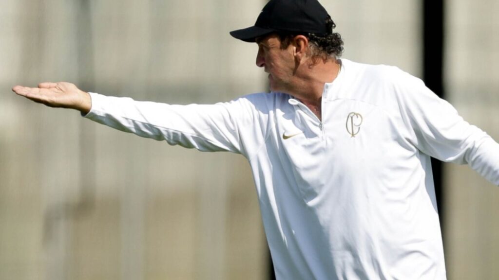 técnico Cuca, enquanto comandava o Corinthians. Ele está de agasalho branco com o escudo do clube e um boné preto, com o braço direito erguido e a palma da mão para cima, apontando algo em orientação no treinamento