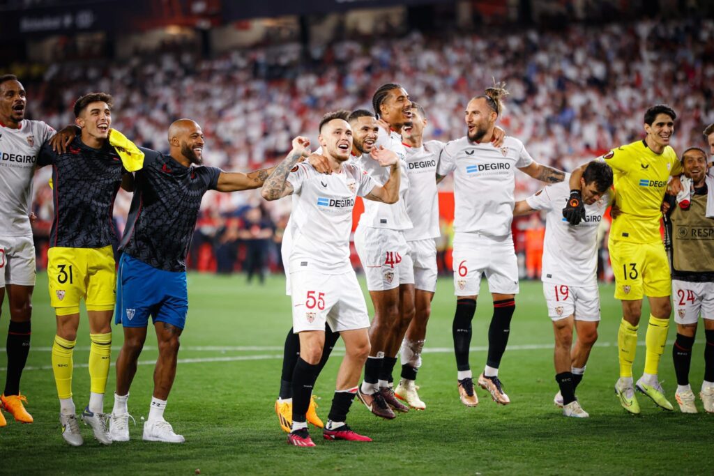 Sevilla vence Manchester United. Jogadores aparecem abraçados comemorando a classificação. Eles aparecem com uniforme branco com detalhes em vermelho.