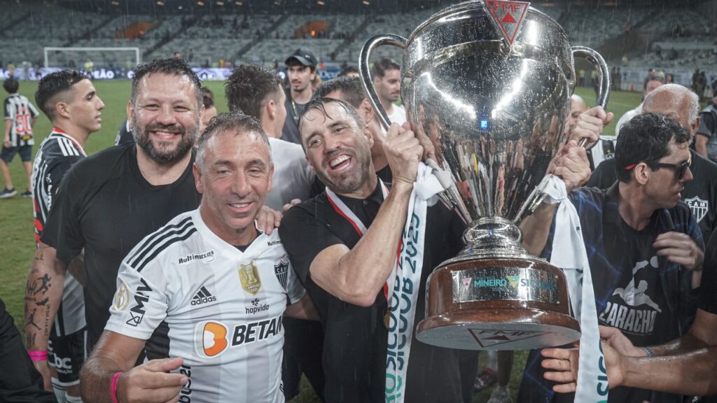 Coudet fica no Atlético-MG: Presidente avalia conduta do treinador após desabafos em coletiva - Eduardo Coudet, técnico do Atlético-MG, ergue a taça do Campeonato Mineiro 2023 e é cercado por outras pessoas vestindo a camisa do Atlético-MG