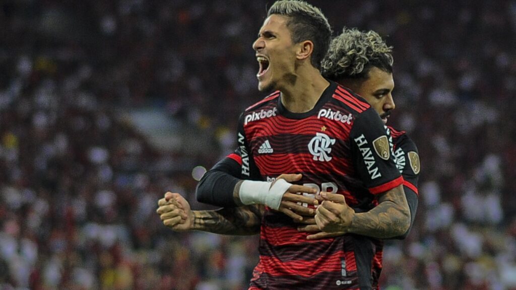 Pedro desfalca o Flamengo: Dois jogadores aparecem se abraçando. Pedro está em mais evidencia e aparece com os braços levantados e gritando. Gabigol está o abraçando de costas.