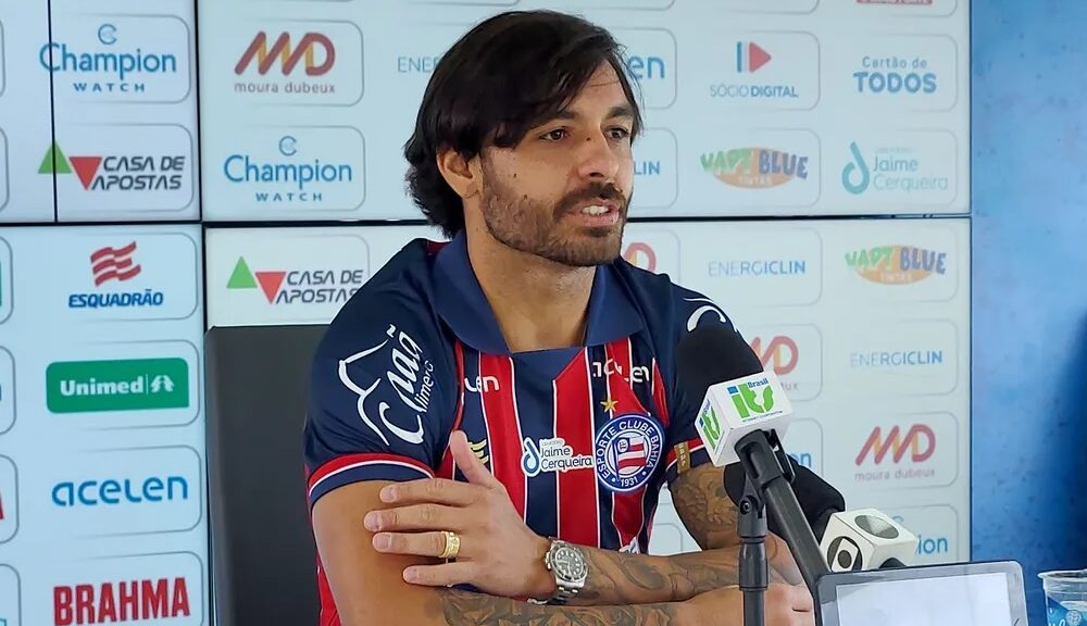 Jogador está sentado diante a um microfone. Ele está dando entrevista coletiva e veste camisa do Bahia, azul com detalhes em vermelho e branco.