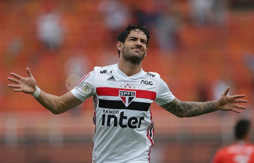 Recuperação de Pato. Jogador aparece no primeiro plano da imagem. Ele está com os braços abertos e veste camiseta branca com detalhes em vermelho e preto.