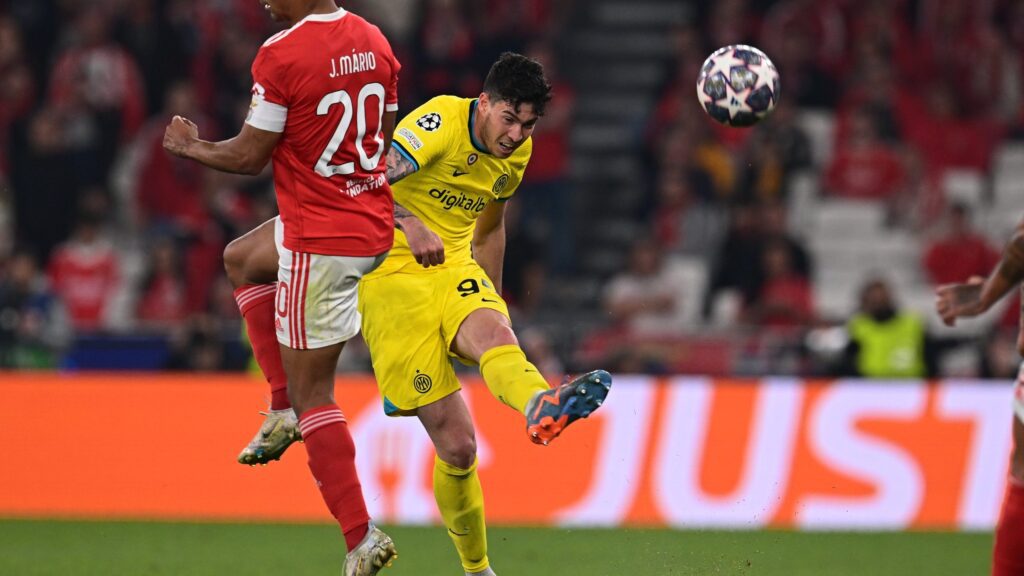 Inter de Milão vence Benfica. Dois jogadores estão no plano da imagem. Um está com uniforme vermelho com detalhes em branco. O outro está com uniforme amarelo com detalhes pretos e chuta a bola alta.