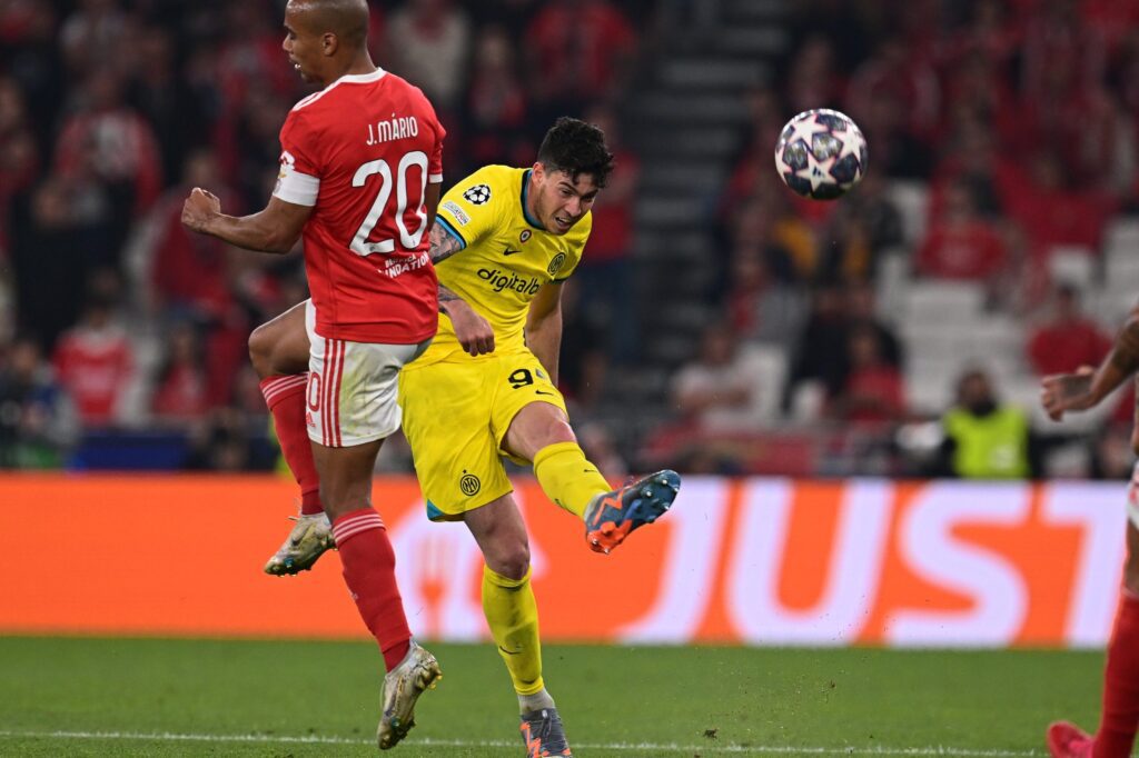 Inter de Milão vence Benfica. Dois jogadores estão no plano da imagem. Um está com uniforme vermelho com detalhes em branco. O outro está com uniforme amarelo com detalhes pretos e chuta a bola alta.