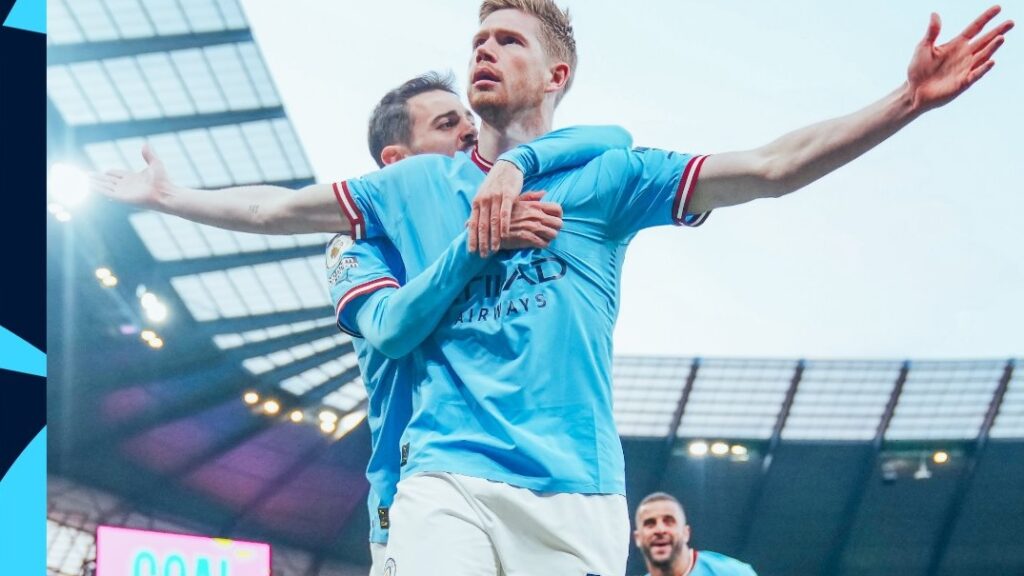 Manchester City vence Arsenal: jogador aparece com os braços abertos comemorando um gol. Ele está sendo abraçado por outro jogador por trás.