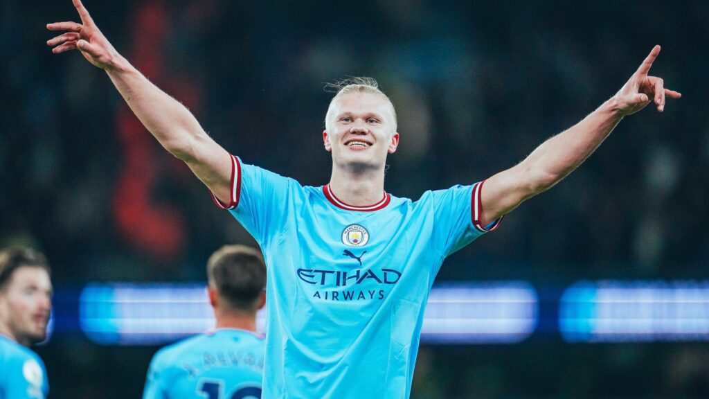 Manchester City é líder da Premier League. Jogador aparece no centro da imagem. Ele está com braços abertos e sorrindo em comemoração ao gol.